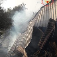 Immagine di un incendio: fumo denso tra crolli di legno e strutture metalliche, scenario caotico e fiamme inesistenti visibili.