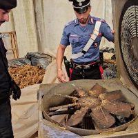 Poliziotto sorveglia una ventola di refrigerazione industriale in ambiente di lavoro, con strumenti e materiali sullo sfondo.