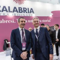 Due uomini in abiti formali si salutano in un ambiente fieristico, con sfondo rosa e logo Calabria in evidenza, atmosfera professionale.