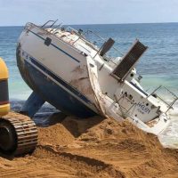 Imbarcazione sommersa in spiaggia, vela rotta e onde sullo sfondo, sabbia e mare azzurro, scenografia marinaresca pericolo e abbandono.