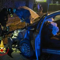 Incidente automobilistico notturno con finestra rotta, soccorritori e veicolo di fronte gravemente danneggiato, luci blu lampeggianti, scena di emergenza.