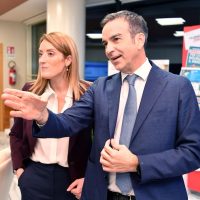 Due persone in abiti formali discutono in un ambiente d'ufficio o evento, con espressioni coinvolte e gesti descrittivi.