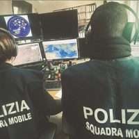 Dipendenti della polizia in centro operativo, con giubbotti ‘POLIZIA SQUADRA MOBILE’ mentre monitorano le mappe e i display.