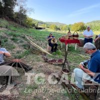gruppo in campagna davanti a un fuoco e strumenti, contesto rurale con colline, persone anziane in abiti casual, attività all’aperto