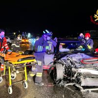 Incidente stradale notturno tra auto danneggiate e soccorritori in divisa fluorescenti, veicoli e barelle su strada.