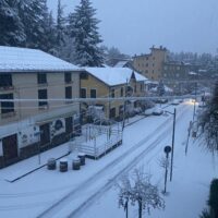 Veduta di una strada cittadina innevata, palazzi gialli ai lati, alberi e lampioni coperti di neve, atmosfera serena invernale.