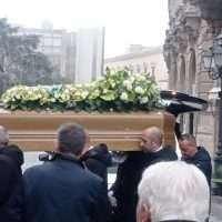 Sarcofago contornato di fiori gialli trasportato da diverse persone in una cerimonia all’aperto, con monumenti sullo sfondo.