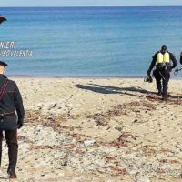 Diversi soccorritori in tuta e giubotto giallo-nero camminano su una spiaggia sabbiosa verso il mare azzurro, in cerca di persone.
