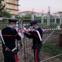Due carabinieri in uniforme bambiniato controllano un lucchetto su una recinzione in un’area verde, con edifici e cantiere sullo sfondo.