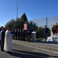 Cerimonia all'aperto con persone in divisa, sacerdote e croce, in un contesto di commemorazione lungo una strada, con parete metallica.