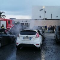 Auto bianca danneggiata in mezzo alla strada, fumo proveniente dal vano motore, vigili del fuoco e curiosi sullo sfondo.