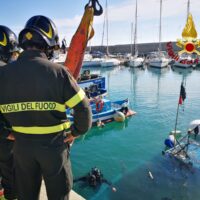 Manovra di soccorso in porto: vigili del fuoco in tuta, piattaforma e gommoni vicino a barche ancorate. Operatori coordinano successivo recupero.