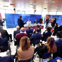 Evento in sala conferenze: pubblico seduto ascolta relatori al tavolo principale, decorazioni blu, atmosfera professionale.