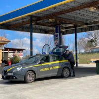 Auto sportiva nera con scritte gialle 'Pine Express' a margine giallo, presente a un distributore di carburante all'aperto in clima soleggiato.