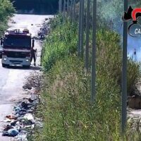 Auto nera parcheggiata vicino a una strada sporca, aiuole di erba alta e rifiuti sul bordo; palina rossa vicina e cartello segnaletico sullo sfondo.