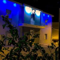 Casa moderna illuminata di blu di notte, luci colorate sulle pareti e cespugli in primo piano.