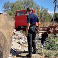 Poliziotto o operatore di sicurezza osserva un'area di cantiere/Rimessa: veicolo rosso, detriti, ruote e cespugli sotto un cielo soleggiato.