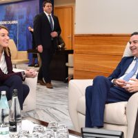 Uomo d'affari in giacca blu sorride mentre parla in una sala riunioni con due colleghi seduti, ambiente professionale e moderno.