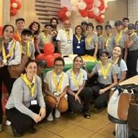 Gruppo di persone in cerimonia, molti con medaglie gialle, sorridono tra palloncini rossi e decorazioni, atmosfera festosa e conviviale.
