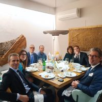 Gruppo di persone in una sala riunioni elegante, sedute attorno a un tavolo con cibo, bevande e una bilancia decorativa sullo sfondo.