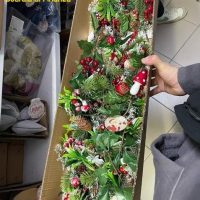 Decorazione natalizia in una scatola di legno con alberello verde, pigne, nastri rossi e luci, maniche di abito visibili a lato.