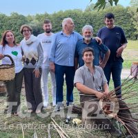 Gruppo di persone in campagna con attrezzi da raccolto, alcuni sorridono, sullo sfondo alberi e strumenti agricoli.