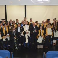 Gruppo di laureandi in abiti accademici, sorridenti, in sala conferenze, periodo cerimonia di laurea, atmosfera festosa.