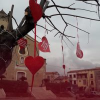 Albero spoglio con cuori rossi pendenti, in centro una piazza e edifici storici sullo sfondo, atmosfera romantica