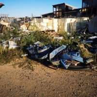 Scena di demolizione e resti metallici arrugginuti in primo piano, case distrutte sullo sfondo e terreno polveroso.