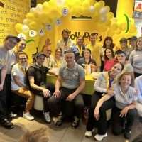 Gruppo di persone sorridenti in un evento promozionale con palloncini gialli sullo sfondo, atmosfera festosa e logo sullo stand.