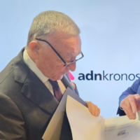 Uomo anziano in giacca e cravatta sfoglia documenti durante una conferenza; sfondo con logo adnkronos.