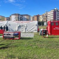 Gruppo di tende e stand informativi in un parco urbano con edifici residenziali sullo sfondo, persone presenti e pannelli colorati.