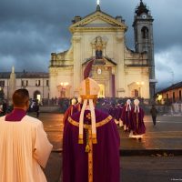 Processione religiosa in piazza davanti a una chiesa barocca, abiti liturgici porpora e illumini rosati al crepuscolo.