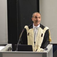 Economista o docente in abito accademico con mantello; parla agli studenti da podio in aula moderna.