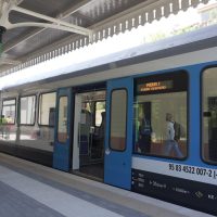 Vagone di una metropolitana blu con porte aperte, in attesa alla pensilina di una stazione, display informative e corridoio di vetro.