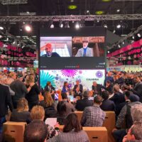 Pubblico di conferenza in sala ampia, relatori sul palco con schermo gigante e sovrascritte, atmosfera vivace e luminosità.