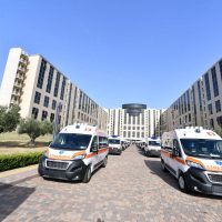 Due ambulanze bianche con strisce arancioni in primo piano, parcheggiate su una strada cipriata, tra edifici moderni e cielo sereno.