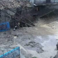 Scena alluvionale in cortile con acqua fangosa, crepe e recinti blu, edificio degradato sullo sfondo.