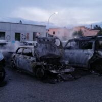 Auto incendiate e carros; scena di veicoli bruciati in un'area urbana, fumo e distruzione visibile sullo sfondo.
