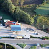 Vista aerea di un piccolo complesso commerciale con tetto blu, parcheggio e strade circostanti tra verdeggiante paesaggio rurale.