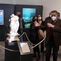 Gruppo di visitatori in una mostra osservano una statua bianca in una vetrina, con viso coperto da mascherina.