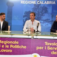 Tre relatori in conferenza, sfondo blu con testo 'Regione Calabria' e pannelli viola, discussione su politiche regionali e servizi.