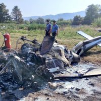 Scena di un incidente: rottami metalici sparsi, due persone in piedi osservano i resti su un campo, veicolo distrutto e montagne sullo sfondo.